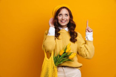 Uzun kırmızı kazaklı ve bereli gülümseyen genç bir kadın elinde lale buketi, üzerinde meyveler olan sarı duvar arkasını gösteren bir çanta taşıyor.