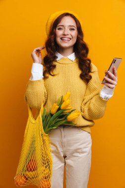 Uzun kırmızı kazaklı ve bereli gülümseyen genç bir kadın elinde lale buketiyle ayakta duruyor. Sarı duvarda meyveler taşıyan, cep telefonu tutan bir çanta taşıyor.