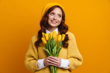 Sırıtkan, uzun kırmızı saçlı, kazaklı ve bereli genç bir kadın. Sarı arka planda lale buketi tutuyor.