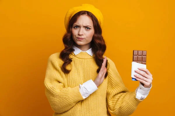 Frowning upset young woman with long red hair in sweater and beret standing holding chocolate bar showing stop gesture over yellow wall background