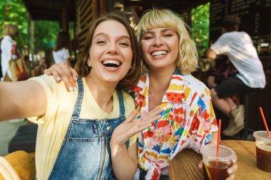 Soda içerken açık havada selfie çeken ve gülen iki kadının görüntüsü.