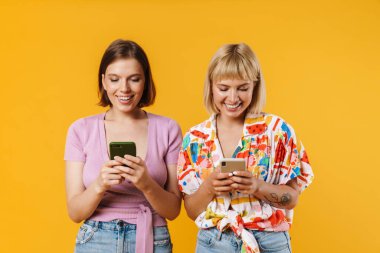 Sarı arka planda duran, ellerinde cep telefonu olan, yaz kıyafetleri giyen iki güzel kız arkadaşın portresi.