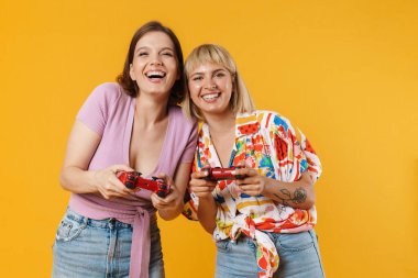 Sarı arka planda izole edilmiş, ellerinde joysticklerle duran, yaz kıyafetleri giyen iki güzel kız arkadaşın portresi.