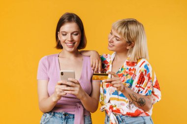 Sarı arka planda izole edilmiş vaziyette duran, elinde cep telefonu olan, kredi kartıyla internetten alışveriş yapan iki güzel kız arkadaşın portresi.