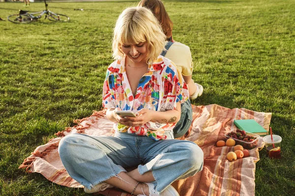 Gülümseyen iki güzel kadının yaz parkında çimlerin üzerinde dinlenirken cep telefonu kullanması.