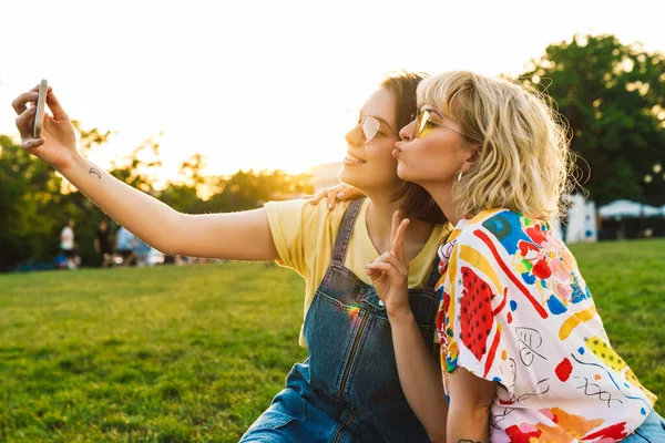 夏の公園で休んでいる間 面白い2人の女性が平和サインをジェスチャーし スマートフォンで自撮り — ストック写真