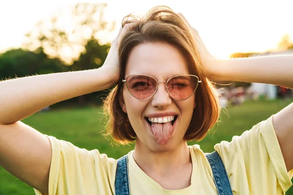 Image of happy brunette woman showing her tongue and winking while resting in summer park