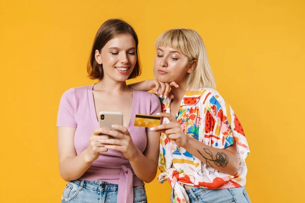 黄色の背景に隔離された立って夏服を着て2人の陽気なガールフレンドの肖像画 携帯電話を保持し クレジットカードでオンラインショッピング — ストック写真