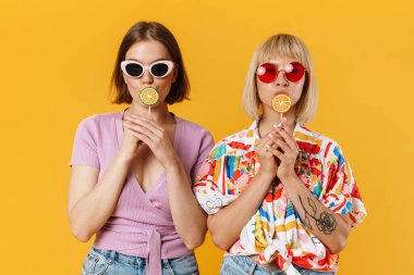 Sarı arka planda izole edilmiş, ellerinde lolipoplar tutan, yaz kıyafetleri giyen iki güzel kız arkadaşın portresi.
