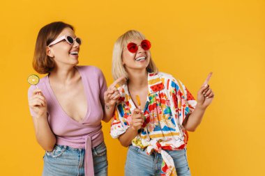 Sarı arka planda izole edilmiş vaziyette duran, elinde lolipoplar olan ve fotokopi çeken iki güzel kız arkadaşın portresi.