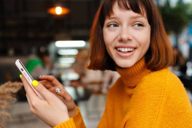 Kafede otururken gülümseyen ve cep telefonu kullanan mutlu kızıl kız.