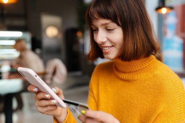 Kafede otururken cep telefonu ve kredi kartı kullanan mutlu bir kızıl kız.