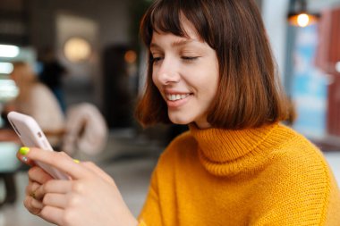 Kafede otururken gülümseyen ve cep telefonu kullanan mutlu kızıl kız.