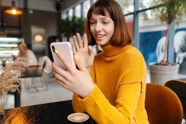 Счастливая Рыжая Девушка Машет Рукой Использует Мобильный Телефон Сидя Кафе — стоковое фото