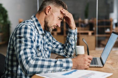 Düşünceli gri adam ofiste otururken dizüstü bilgisayarla çalışıyor.