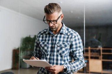 Ciddi, beyaz saçlı bir adam ofiste dururken evraklarla çalışıyor.