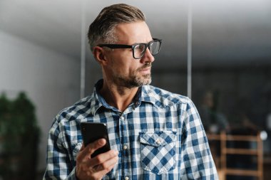 Ofiste cep telefonu kullanan gözlüklü, ciddi, beyaz saçlı bir adam.