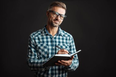 Gri duvar arkaplanından izole edilmiş not defterine notlar yazan pozitif gri saçlı bir adamın resmi