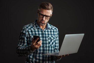 Gri saçlı şok olmuş adamın görüntüsü, dizüstü bilgisayar ve cep telefonu kullanarak gri duvar arkaplanı üzerinde izole edilmiş.