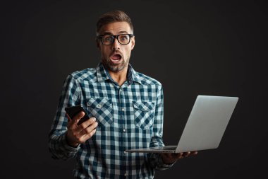 Gri saçlı şok olmuş adamın görüntüsü, dizüstü bilgisayar ve cep telefonu kullanarak gri duvar arkaplanı üzerinde izole edilmiş.