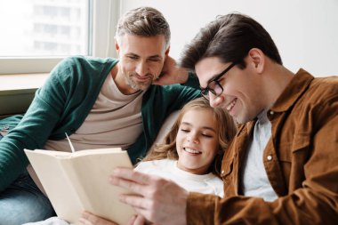 Mutlu eşcinsel ebeveynler evde kızlarıyla birlikte kitap okuyor.
