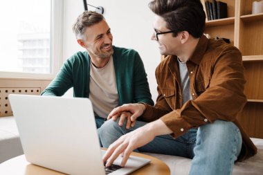 Mutlu genç eşcinsel çift evdeki kanepede otururken dizüstü bilgisayar kullanıyor.