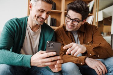 Oturma odasındaki koltukta otururken cep telefonu kullanan mutlu, genç eşcinsel çift.