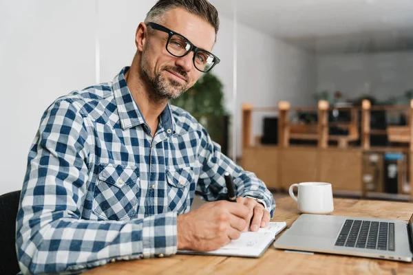 Agradable Hombre Gris Anotando Notas Mientras Trabaja Con Ordenador Portátil — Foto de Stock