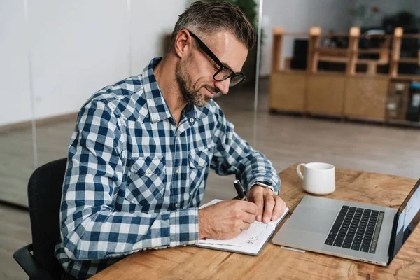 Agradable Hombre Gris Anotando Notas Mientras Trabaja Con Ordenador Portátil — Foto de Stock