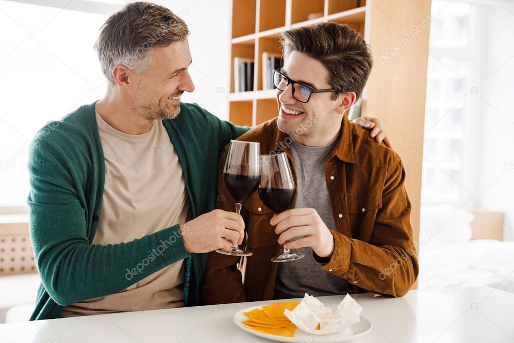 Happy caucasian gay couple hugging while drinking wine at home