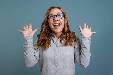 Gözlüklü heyecanlı kız gülümserken elleri yukarıda mavi arka planda poz veriyor.