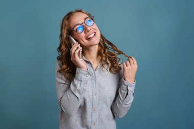 Mavi arka planda izole edilmiş cep telefonuyla konuşurken gülümseyen gözlüklü mutlu kız.