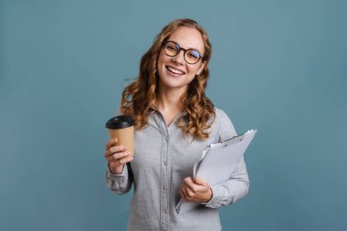 Gülümseyen çekici genç kadın elinde not defteri ve kahve fincanıyla mavi arka planda izole bir şekilde duruyor.