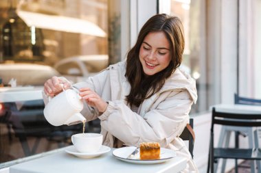 Neşeli genç bayan, açık kafede kekle çay içiyor.