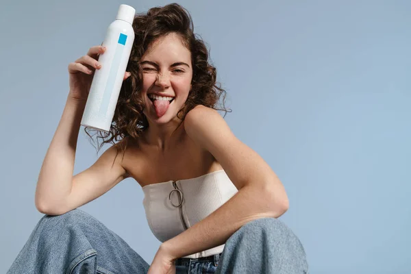 Joyful curly girl showing thermal water and her tongue isolated over blue background