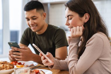 Gülümseyen bir çiftin portresi akıllı telefonlar kullanıyor ve evde rahat bir mutfakta kahvaltı ederken konuşuyor.