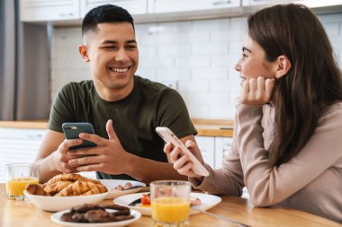 Gülümseyen bir çiftin portresi akıllı telefonlar kullanıyor ve evde rahat bir mutfakta kahvaltı ederken konuşuyor.
