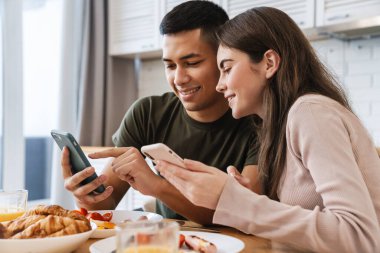 Mutfakta lezzetli bir kahvaltı yaparken cep telefonu kullanan mutlu ve genç bir çift.