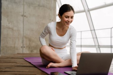 Evde yoga yaparken dizüstü bilgisayar kullanan gülümseyen genç bir kadın.