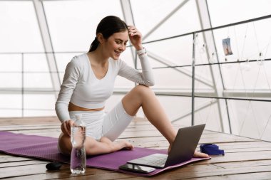 Evde yoga yaparken dizüstü bilgisayar kullanan esmer bir kadın.