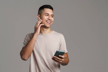 Gri duvarda izole edilmiş cep telefonu kullanırken kulaklıklı mutlu esmer adam gülümsüyor.