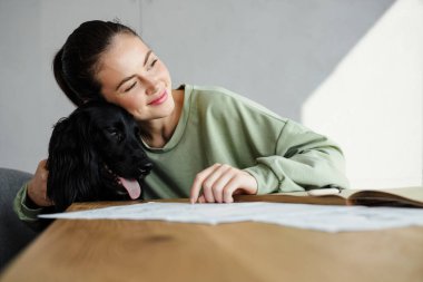 Mutlu esmer kadın evde ders çalışırken, masada otururken köpeğini okşuyor.