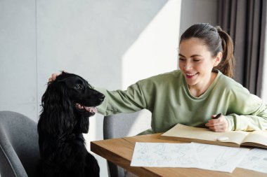 Mutlu esmer kadın evde ders çalışırken, masada otururken köpeğini okşuyor.