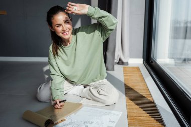 Gülümseyen genç kadın tasarımcı pencerenin önünde otururken çizimler çiziyor, güneşten korunuyor.