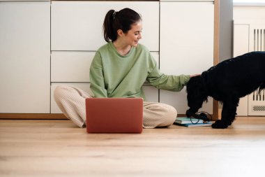 Genç, mutlu, esmer bir kadın köpeğiyle yerde otururken dizüstü bilgisayar kullanıyor.