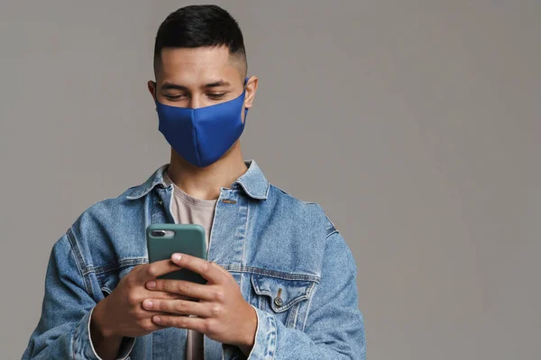 Brunett Ung Nöjd Man Ansiktsmask Med Mobiltelefon Isolerad Över Grå — Stockfoto