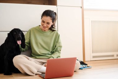 Genç, mutlu, esmer bir kadın köpeğiyle yerde otururken dizüstü bilgisayar kullanıyor.