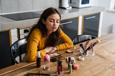 Genç esmer kadın mutfakta otururken makyaj yapıyor.