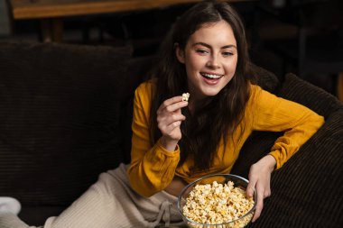 Evdeki kanepede oturmuş patlamış mısır yiyen genç bir kadın.