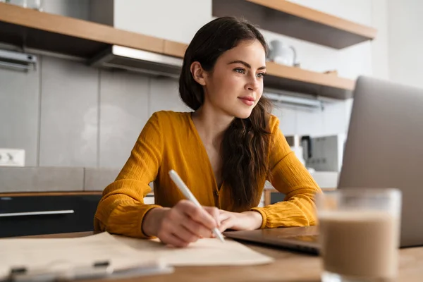 Joven Mujer Complacida Escribiendo Notas Mientras Trabaja Con Ordenador Portátil —  Fotos de Stock